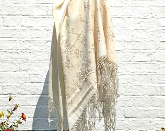 May include: A cream-colored shawl featuring an intricate embroidered pattern of small squares and floral motifs. The garment has a draped neckline and long, flowing fringe trim along the edges, creating a textured appearance throughout the fabric.