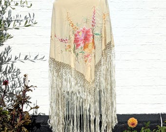 May include: A cream-colored triangular shawl featuring an embroidered floral design in shades of pink, orange, and green. The shawl is finished with a wide, knotted macramé border and long, flowing fringe along the two bottom edges.