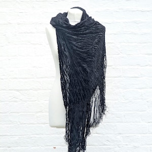 May include: A black scarf featuring a textured, crinkled fabric surface and long, delicate fringe detailing along the edges. The scarf is draped over a mannequin, showcasing its lightweight material and the intricate pattern of the fabric weave.