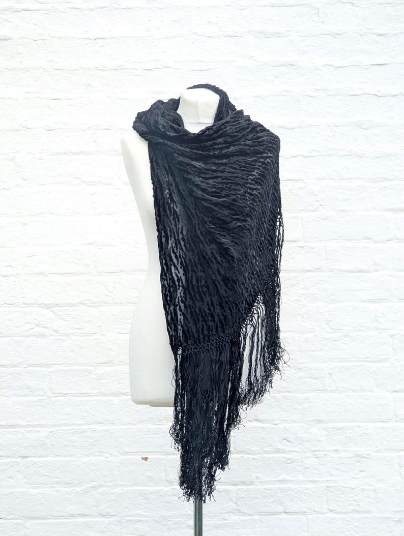 May include: A black scarf featuring a textured, crinkled fabric surface and long, delicate fringe detailing along the edges. The scarf is draped over a mannequin, showcasing its lightweight material and the intricate pattern of the fabric weave.