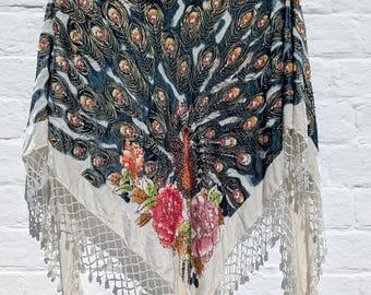 May include: A velvet shawl with a peacock feather design in teal, gold, and orange. The shawl has a cream-colored border with a lace fringe and a floral design at the bottom.