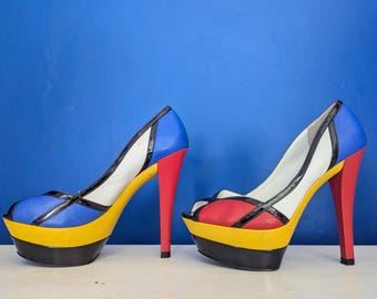 Puede incluir: Un par de zapatos de tacón alto coloridos. Los zapatos presentan una puntera azul, un tacón rojo y una plataforma amarilla y negra. La parte superior es blanca con ribete negro. Los tacones miden unos 10 cm de alto.