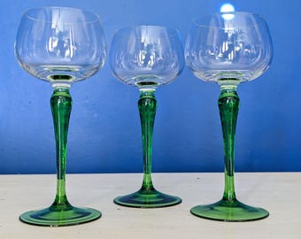 May include: Three clear wine glasses with green stems and bases. The glasses have a rounded bowl shape and a long, elegant stem. The green color is a vibrant, rich shade. The glasses are arranged in a row against a blue background.