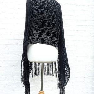 May include: A black shawl featuring a textured, raised velvet-like pattern in a horizontal, wavy design. The fabric is semi-sheer and lightweight, finished with long, thin, matching black fringe trim along the bottom edge of the garment.