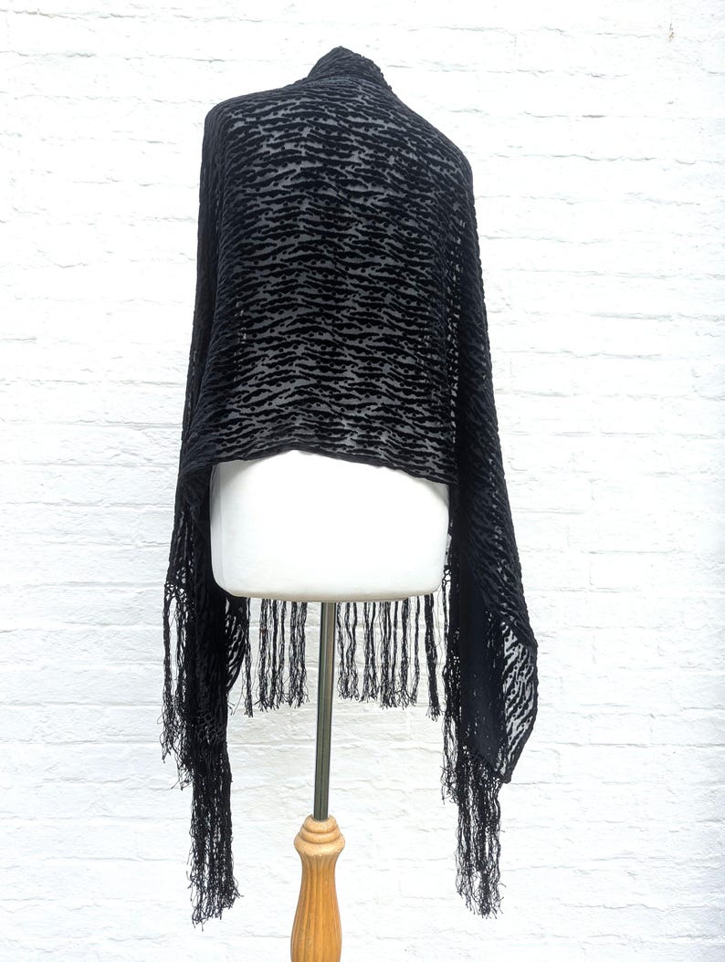 May include: A black shawl featuring a textured, raised velvet-like pattern in a horizontal, wavy design. The fabric is semi-sheer and lightweight, finished with long, thin, matching black fringe trim along the bottom edge of the garment.