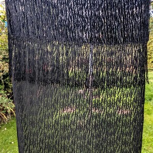 May include: A rectangular black scarf featuring a semi-sheer fabric with a textured, raised velvet-like pattern in a repeating vertical stripe design. The scarf includes a horizontal fringe detail across the middle and a long fringe at the bottom edge.