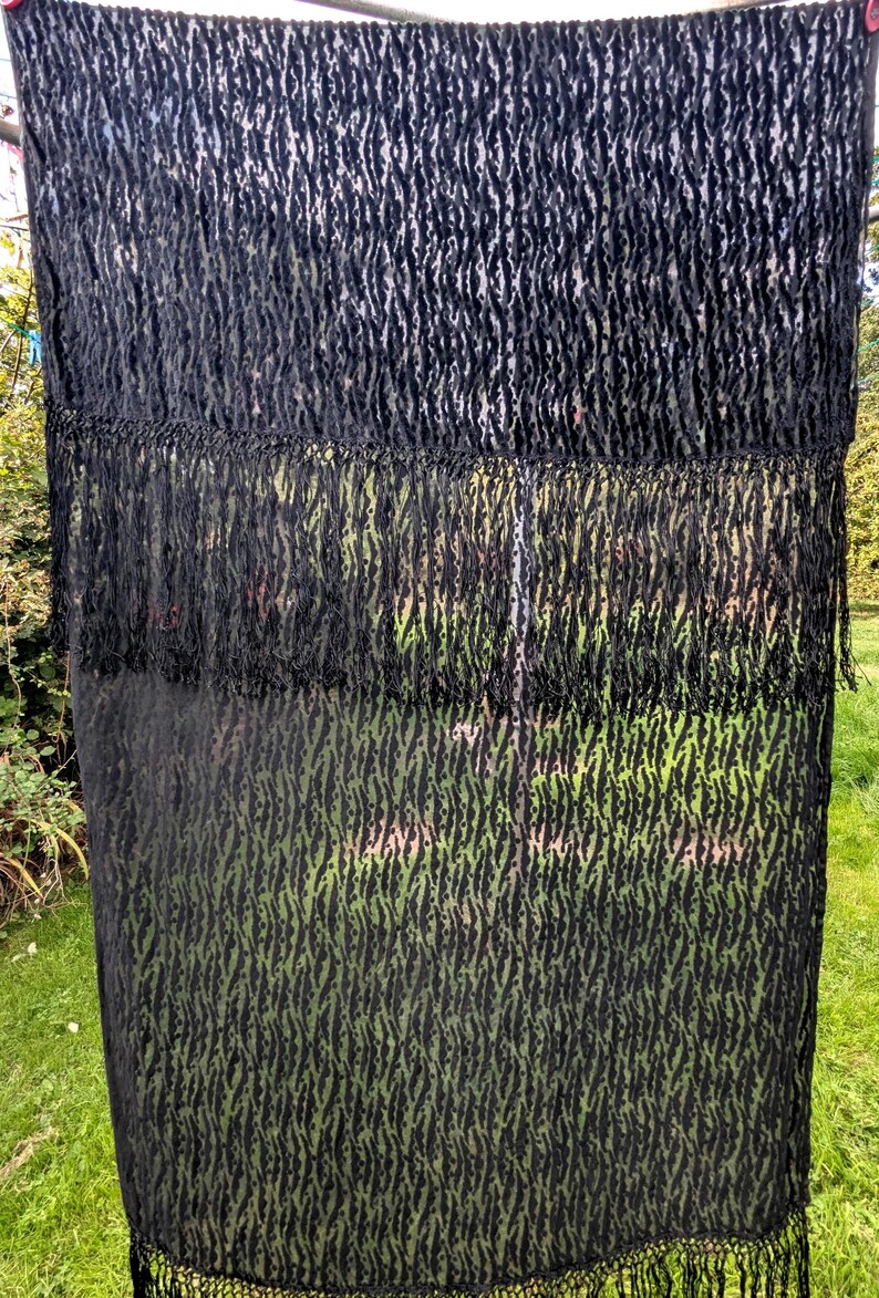 May include: A rectangular black scarf featuring a semi-sheer fabric with a textured, raised velvet-like pattern in a repeating vertical stripe design. The scarf includes a horizontal fringe detail across the middle and a long fringe at the bottom edge.