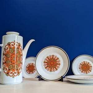 May include: A white ceramic coffee pot and matching plates with an orange and green floral design. The set includes a tall coffee pot, three plates, and a smaller plate. The plates have a brown border and are set against a blue background.