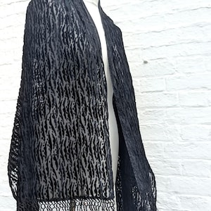 May include: A black velvet shawl featuring a textured, repeating vertical pattern of raised stripes and dots. The rectangular garment is finished with long, thin, matching black fringe along the bottom edge, draping over a mannequin form.