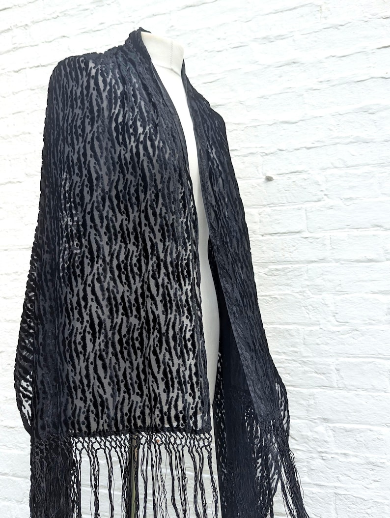 May include: A black velvet shawl featuring a textured, repeating vertical pattern of raised stripes and dots. The rectangular garment is finished with long, thin, matching black fringe along the bottom edge, draping over a mannequin form.