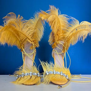 May include: A pair of yellow high-heeled sandals with feather and rhinestone embellishments. The sandals have a strappy design with a delicate ankle tie. The background is a solid blue.