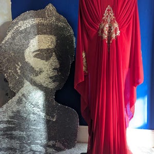 Könnte beinhalten: Ein leuchtend rotes Kleid mit goldener Stickerei auf einer Schaufensterpuppe. Das Kleid hat ein fließendes Design und wird vor einer blauen Wand ausgestellt. Links ist ein paillettenbesetztes Porträt.