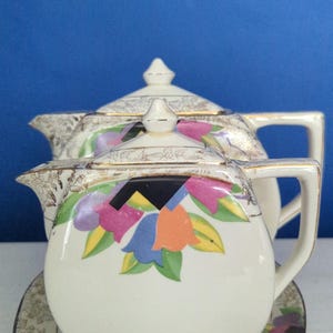 May include: Cream-coloured teapot with a matching lid and saucer. The teapot features a colourful floral design with gold trim. The background is a solid blue.