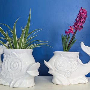 Puede incluir: Dos macetas de cerámica blanca en forma de pez, una con una planta verde y la otra con una flor de jacinto rosa. Las macetas tienen un diseño detallado y en espiral.