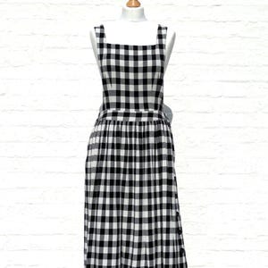 May include: A sleeveless midi dress featuring a black and white gingham check pattern. The dress has a square neckline, a fitted bodice with a defined waistline, and a pleated skirt. A row of black buttons runs vertically along the side seam.