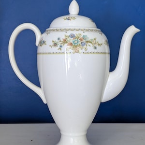 May include: White ceramic teapot with a floral design. The teapot has a tall, slender body, a curved handle, and a long spout. The lid is topped with a small finial. The floral pattern includes blue, orange, and green flowers. The teapot sits on a pedestal base.