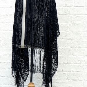 May include: A black shawl featuring a semi-sheer burnout velvet texture with a vertical abstract pattern. The garment has an open front design and is finished with long, thin, matching black fringe trim along the bottom hemline.