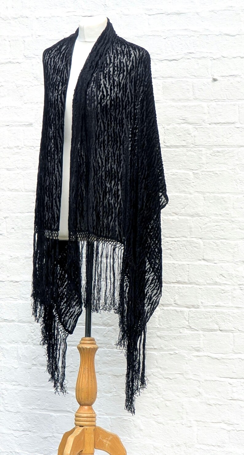 May include: A black shawl featuring a semi-sheer burnout velvet texture with a vertical abstract pattern. The garment has an open front design and is finished with long, thin, matching black fringe trim along the bottom hemline.