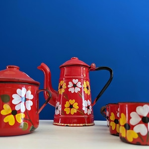 Puede incluir: Un juego de té de esmalte rojo con una tetera, una olla con tapa y dos tazas. El juego está decorado con diseños florales blancos y amarillos. La tetera tiene un asa y un pico negros. El fondo es azul liso.