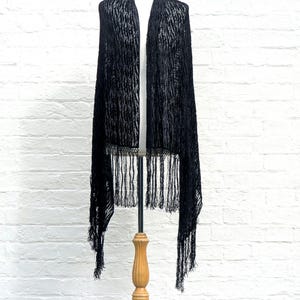 May include: A long black shawl featuring a textured, semi-sheer fabric with a subtle vertical pattern. The garment is draped over a mannequin and finished with long, thin fringe detailing along the bottom edge, creating a flowing silhouette.
