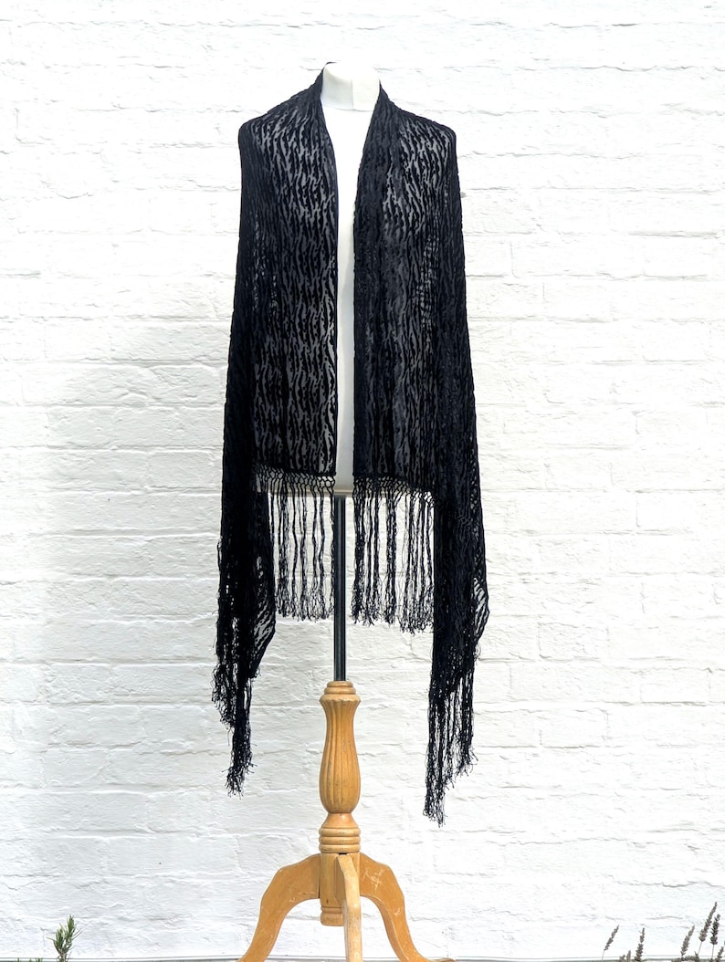 May include: A long black shawl featuring a textured, semi-sheer fabric with a subtle vertical pattern. The garment is draped over a mannequin and finished with long, thin fringe detailing along the bottom edge, creating a flowing silhouette.