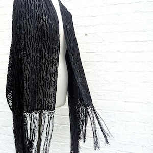 May include: A black shawl featuring a textured, raised velvet-like pattern against a sheer base. The garment is rectangular in shape and finished with long, thin, knotted fringe detailing along the bottom edges, creating a draped appearance.