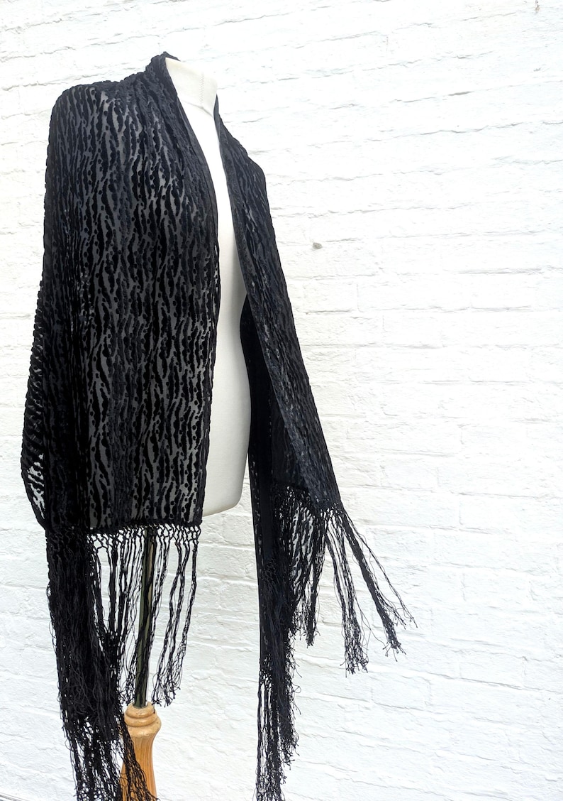 May include: A black shawl featuring a textured, raised velvet-like pattern against a sheer base. The garment is rectangular in shape and finished with long, thin, knotted fringe detailing along the bottom edges, creating a draped appearance.