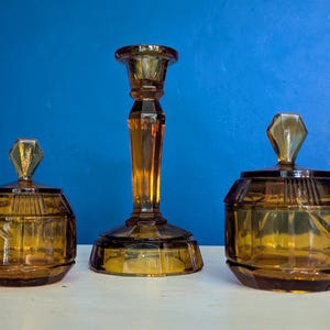Könnte beinhalten: Ein Set aus bernsteinfarbenen Glasdekorationsartikeln. Das Set umfasst einen hohen Kerzenhalter und zwei passende Gläser mit Deckel. Das Glas hat ein geometrisches Design und rautenförmige Knäufe auf den Deckeln. Vintage-Stil.