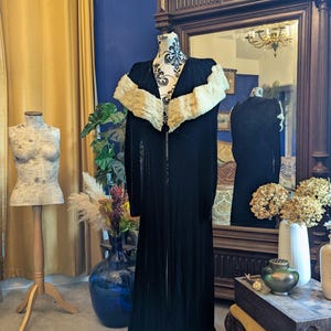 Puede incluir: Un abrigo largo de terciopelo negro con cuello de piel color crema expuesto en un maniquí. El abrigo es largo hasta el suelo y tiene una parte delantera abotonada. El maniquí está frente a un gran espejo y un armario de madera oscura.