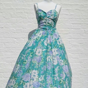 May include: A vintage floral dress in teal, white, and lavender. The dress has a fitted bodice with button details and spaghetti straps, flowing into a full skirt. The dress is displayed on a mannequin. The dress is a classic vintage style.