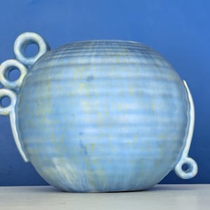 May include: A light blue ceramic vase with a round shape and decorative circular handles. The vase has a textured surface and a light blue colour with hints of yellow. The vase is on a white surface against a blue background.