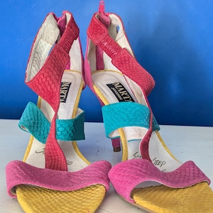 May include: A pair of Marta Jonsson high-heeled sandals featuring a multi-colored snakeskin-textured pattern in pink, red, teal, and yellow. The shoes have a T-strap design, an open toe, and a zipper closure at the back of the heel.