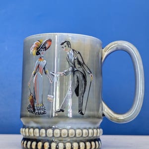 Puede incluir: Taza de cerámica gris con un asa grande, con una ilustración vintage de un hombre y una mujer con ropa formal. La taza tiene una base decorativa con un patrón repetido de pequeñas formas redondeadas. El fondo es azul liso.