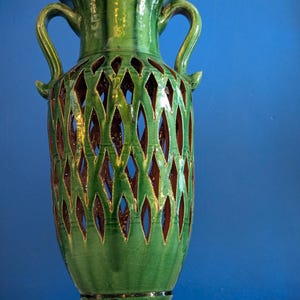 May include: A tall, green ceramic vase with two handles and a flared rim. The vase features a diamond-shaped cut-out design revealing a brown interior. The vase is placed against a blue background.