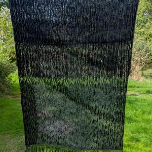 May include: A rectangular black scarf featuring a semi-sheer, textured, and slightly raised pattern throughout the fabric. The scarf is finished with long, thin, black fringe detailing along the bottom edge, creating a consistent dark aesthetic.
