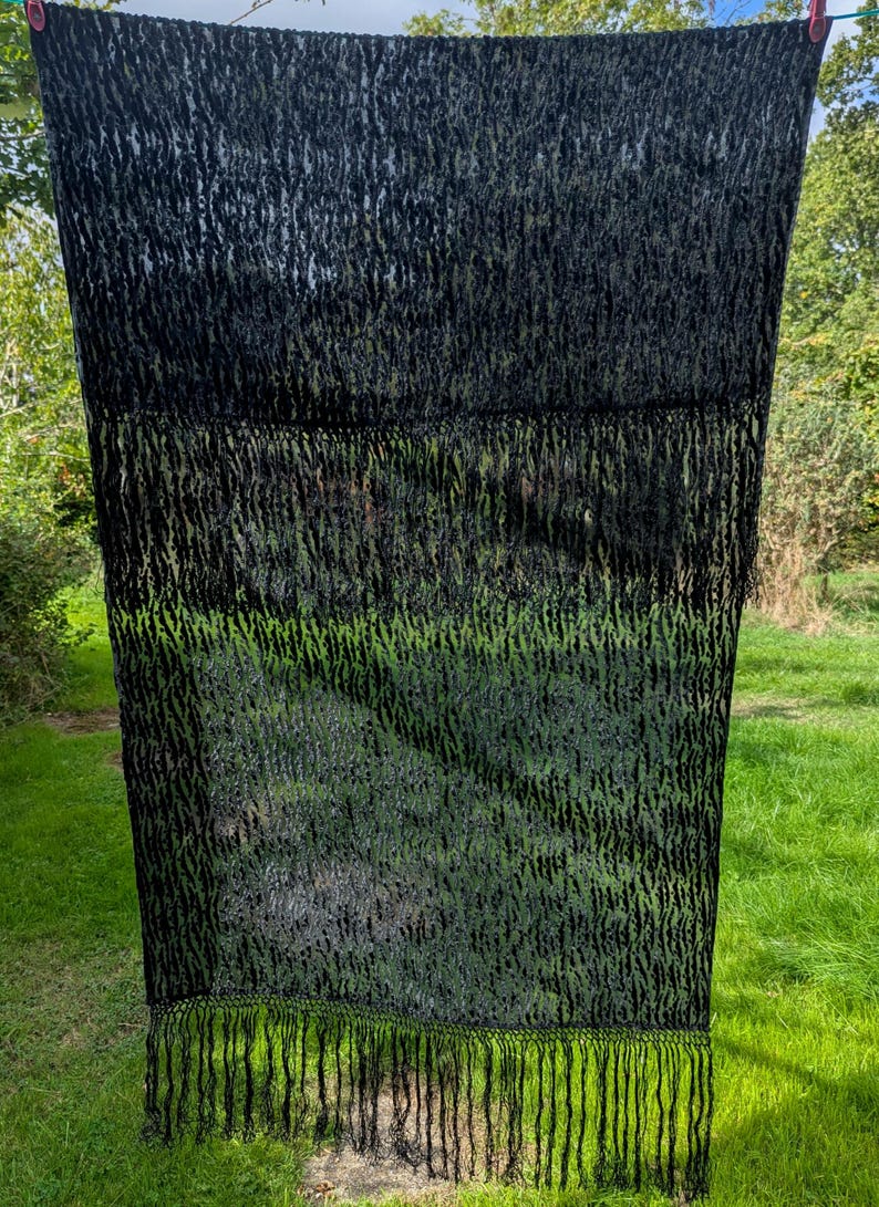May include: A rectangular black scarf featuring a semi-sheer, textured, and slightly raised pattern throughout the fabric. The scarf is finished with long, thin, black fringe detailing along the bottom edge, creating a consistent dark aesthetic.