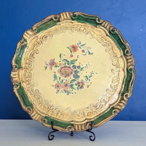 May include: A round, antique serving tray with a scalloped edge. The tray has a cream-coloured centre with a floral design. The rim is gold with green accents. The tray is displayed on a black metal stand.