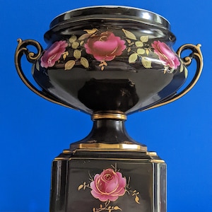 Puede incluir: Una urna de cerámica negra con detalles dorados y diseños florales de rosas rosas. La urna tiene dos asas y se asienta sobre una base cuadrada.