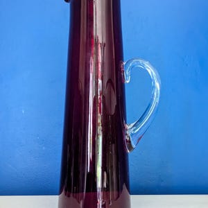 May include: A tall, elegant purple glass pitcher with a clear glass handle. The pitcher has a slender, cylindrical shape and a pouring spout. The handle is curved and attached to the side of the pitcher. The pitcher is likely used for serving beverages.