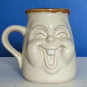 May include: Cream-colored ceramic mug with a smiling face design. The mug features a large, expressive face with closed eyes, rosy cheeks, and a wide, toothy grin. The rim is trimmed with a brown band, and it has a curved handle.