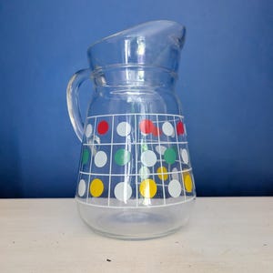 Puede incluir: Una jarra de vidrio transparente con asa y boquilla. La jarra está decorada con un patrón de cuadrícula blanca y lunares de colores en rojo, verde, amarillo y blanco. La jarra está sobre una superficie blanca sobre un fondo azul.