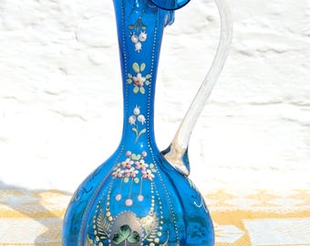 May include: A cobalt blue glass pitcher with a ruffled rim and a clear glass handle. The body features hand-painted floral motifs, including small pink and white flowers, green leaves, and a central green shamrock design surrounded by gold dots.