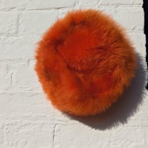 Puede incluir: Un objeto redondo y esponjoso de color naranja brillante, posiblemente un sombrero o un elemento decorativo, se encuentra sobre una pared de ladrillos blancos. El objeto tiene una textura suave y densa, con un color saturado. La iluminación crea una sombra.