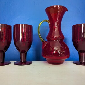 Può includere: Set di quattro bicchieri da bere in vetro rosso, tra cui una caraffa con manico verde. I bicchieri hanno una ciotola arrotondata e uno stelo. La caraffa ha una base larga e un collo stretto.
