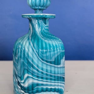 Puede incluir: Una botella de vidrio azul turquesa con vetas blancas y un diseño acanalado. La botella tiene un tapón decorativo azul turquesa.