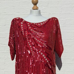 May include: A red sequined top with short sleeves, displayed on a mannequin. The top features a pattern of sequins and small white beads. The neckline is embellished with small white beads. The garment is a vibrant red colour.