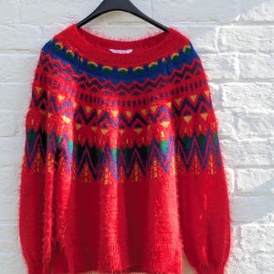 Könnte beinhalten: Roter, flauschiger Pullover mit einem bunten geometrischen Muster. Der Pullover hat lange Ärmel und einen runden Ausschnitt.