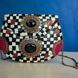 Könnte beinhalten: Eine dekorative Handtasche mit einem schwarz-weißen Schachbrettmuster. Die Tasche hat rote und goldene Akzente, schwarze kreisförmige Steine und einen Kettenriemen. Die Tasche steht auf einer weißen Oberfläche mit blauem Hintergrund.