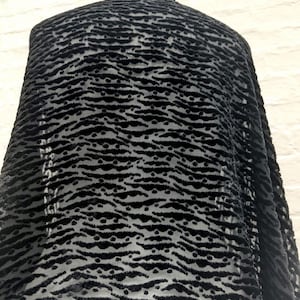 May include: A black fabric featuring a raised, textured velvet pattern of horizontal wavy lines and small dots. The material has a semi-sheer base, creating a contrast between the opaque, fuzzy velvet elements and the translucent background sections.