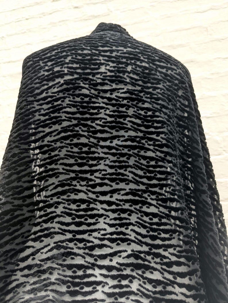 May include: A black fabric featuring a raised, textured velvet pattern of horizontal wavy lines and small dots. The material has a semi-sheer base, creating a contrast between the opaque, fuzzy velvet elements and the translucent background sections.